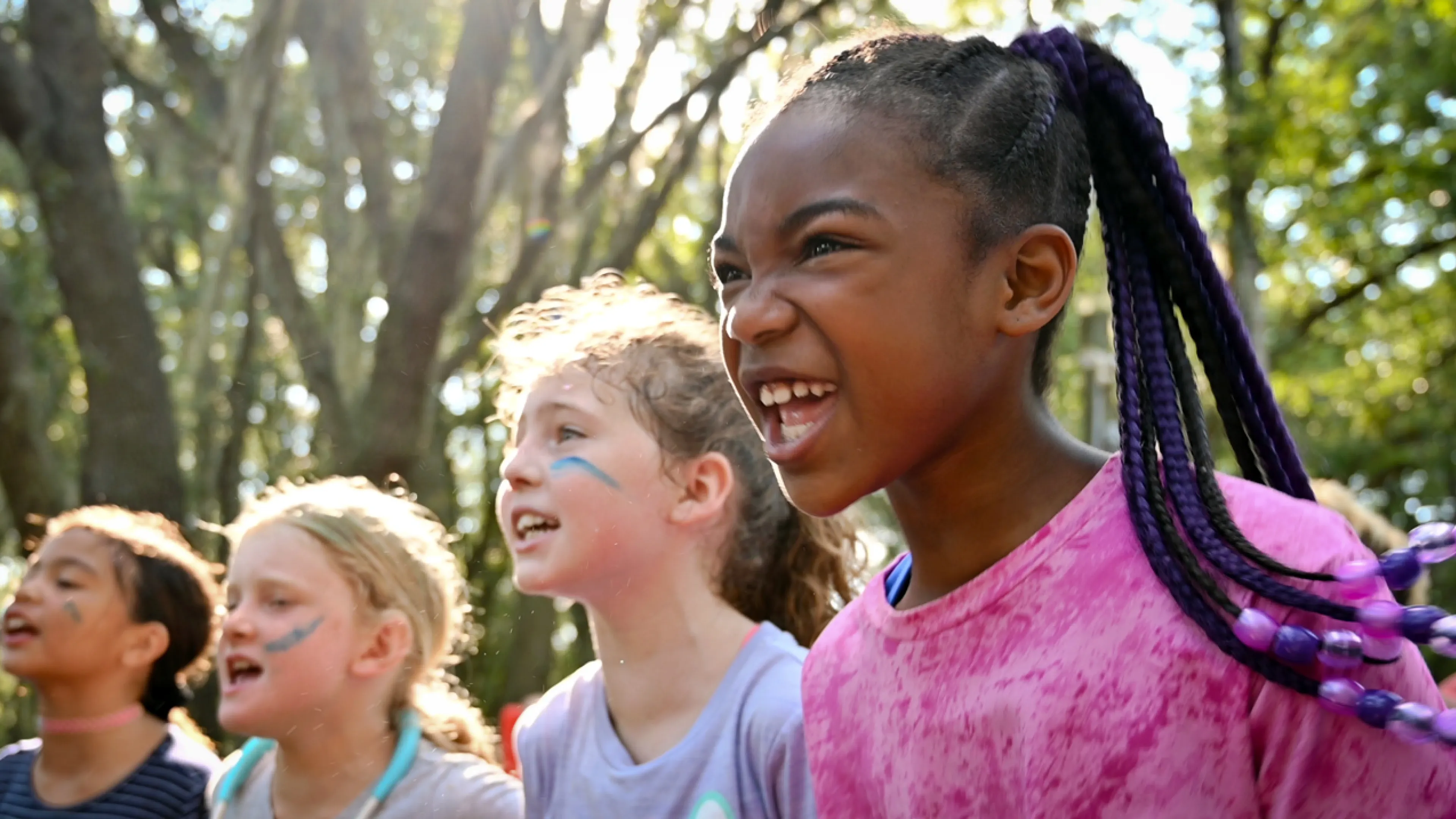 Four school age kids shouting a chant outside at camp. Sun peeks through the trees and shines their heads.